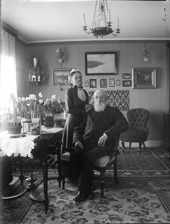 Interiör hos Anna Cegrell och Hans Cederberg på Bergsgatan 50 i Gränna. Mannen med stort vitt skägg och på sitt sista år sitter på en stol med hustrun stående vid sin sida.