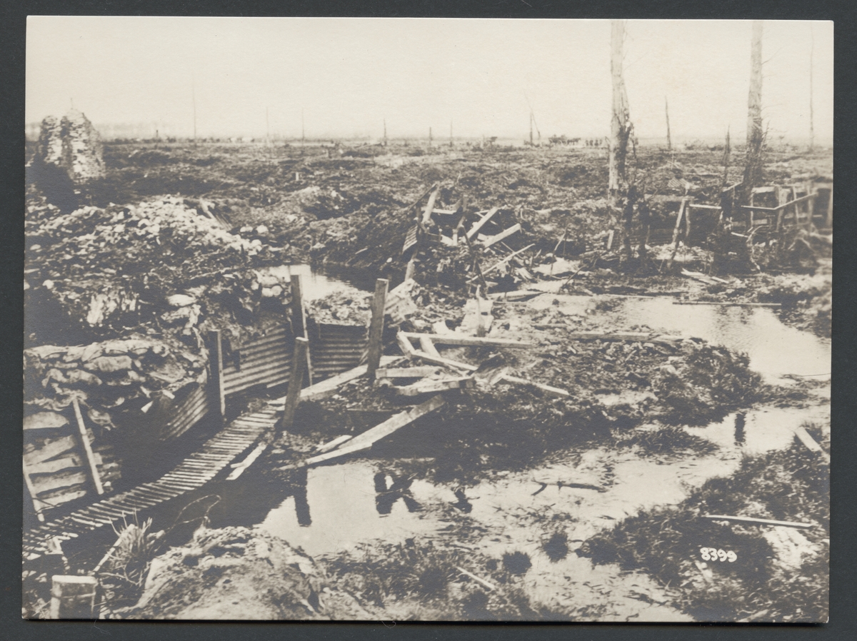 Bilden visar ett slagfält från första världskriget. Området är helt uppgrävt av artillerield och täckt med vatten, slam och spillror av krigsmateriel. I bakgrunden syns ett kolon hästvagnar som passerar på en väg.