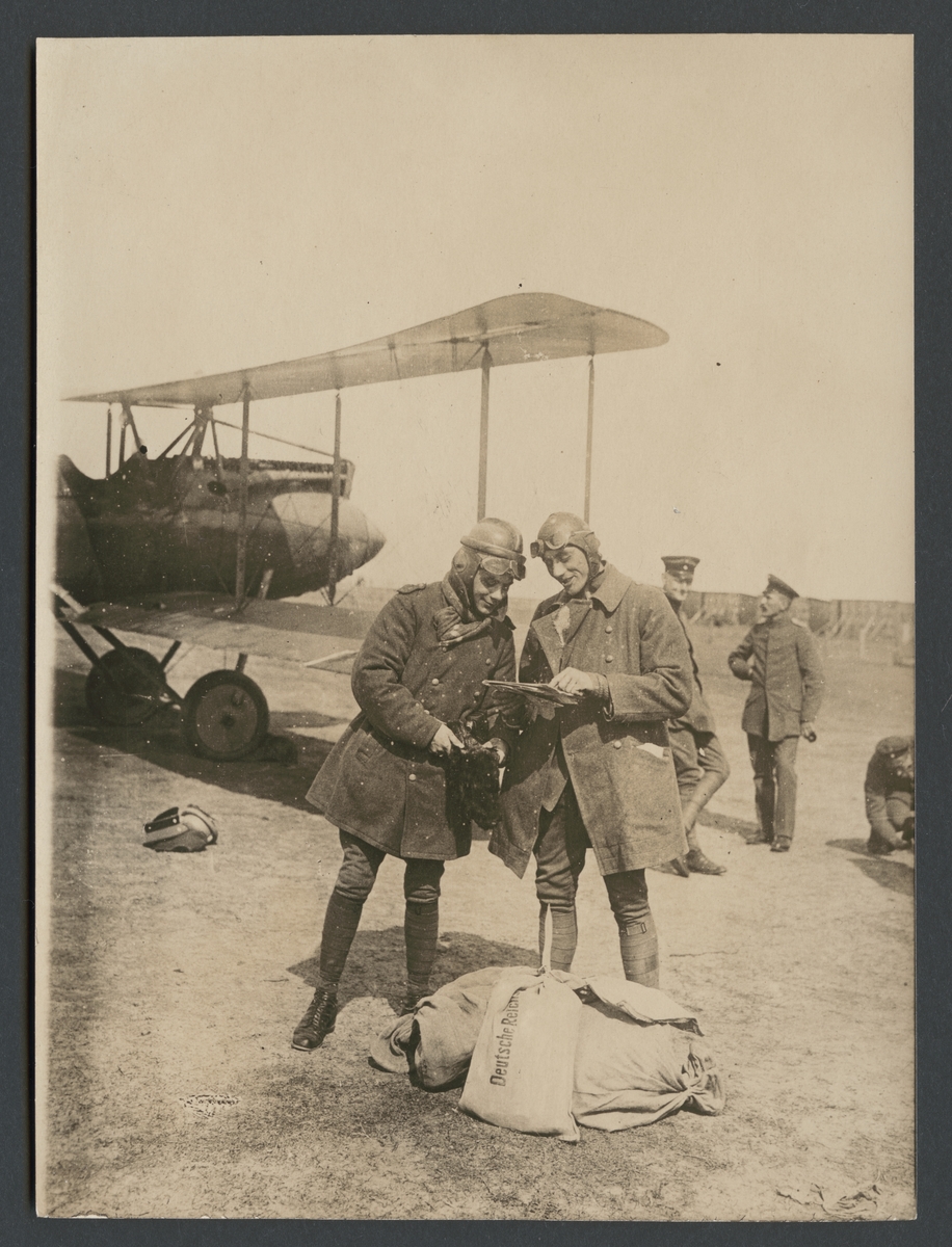 Bilden visar två piloter som titta på en karta. De står framför ett dubbeldäckare och bär flyghuva med glasögon. Till deras fötter ligger postsäckar.

Originaltext: "Flygmaskinens förare och hans ledsagare studera kartan före starten."