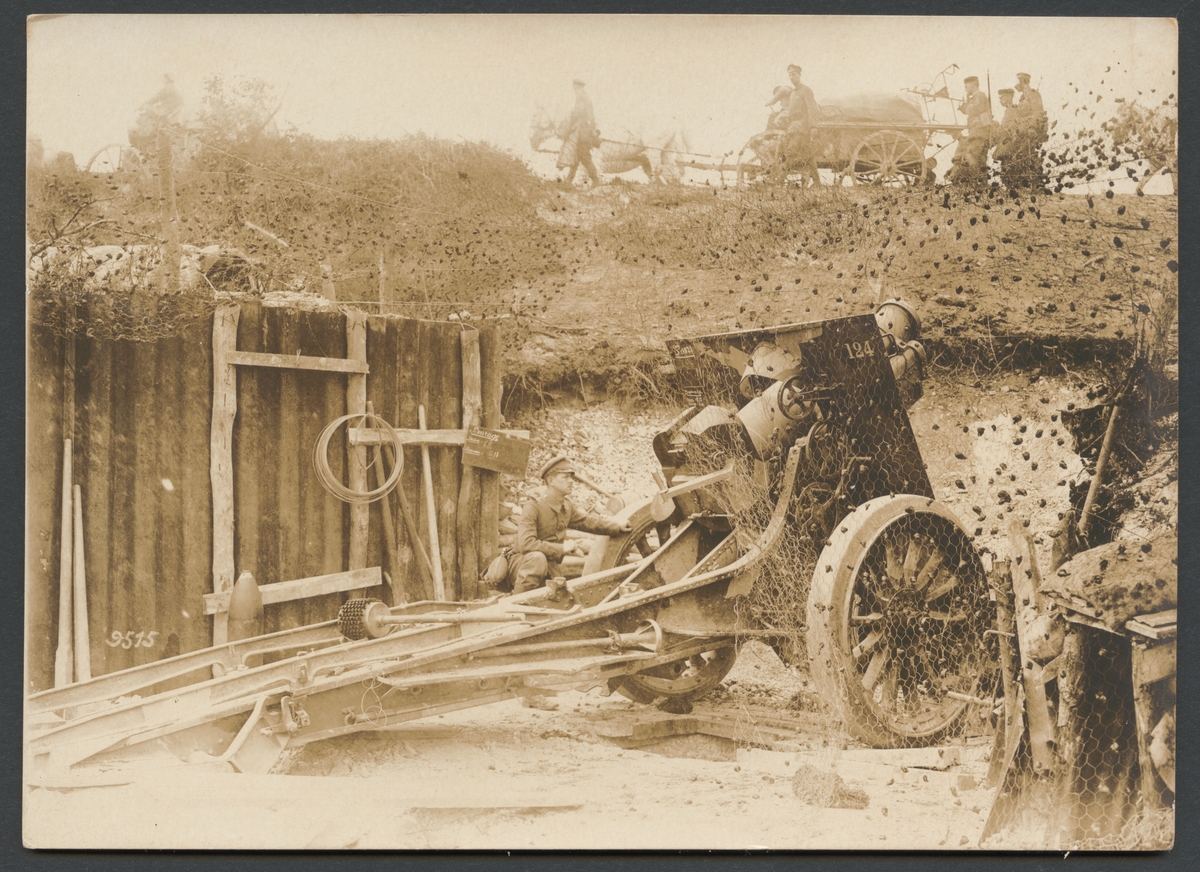 Bilden visar en soldat som undersöker en stor artilleripjäs i en artillriställning som är täckt med en maskeringsnät. I bakgrunden syns en grupp soldater med häst och vagn marscherar förbi.