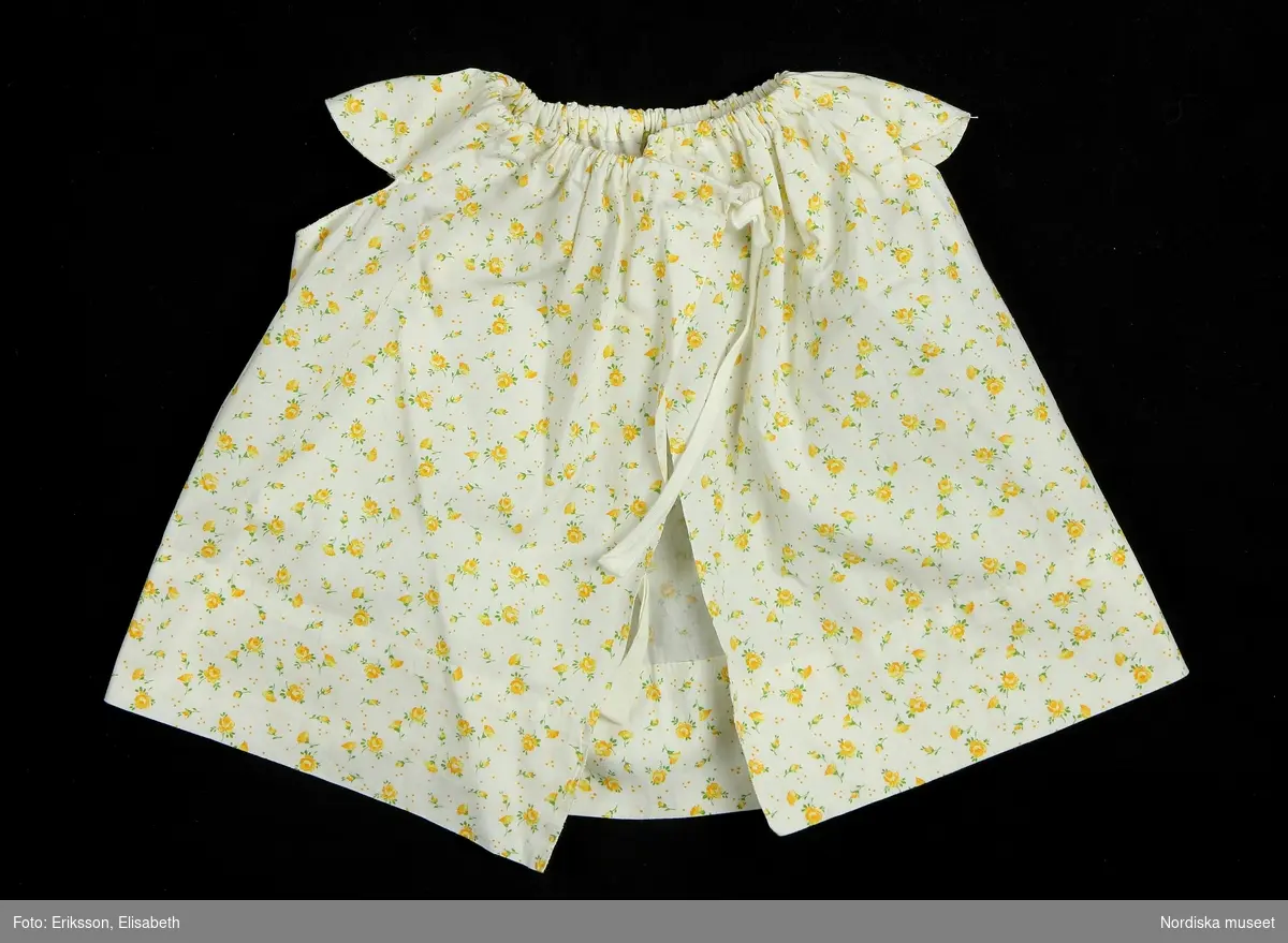 Spädbarnsdräkt i två delar, bestående av klänning med tillhörande trosa. Klänningen är av enkel modell, sydd i ett stycke av småblommigt bomullstyg, med gula rosor på vit botten. Holkärmar, rynkning vid halsen, fållad kant. Knytning bak med bomullsband.