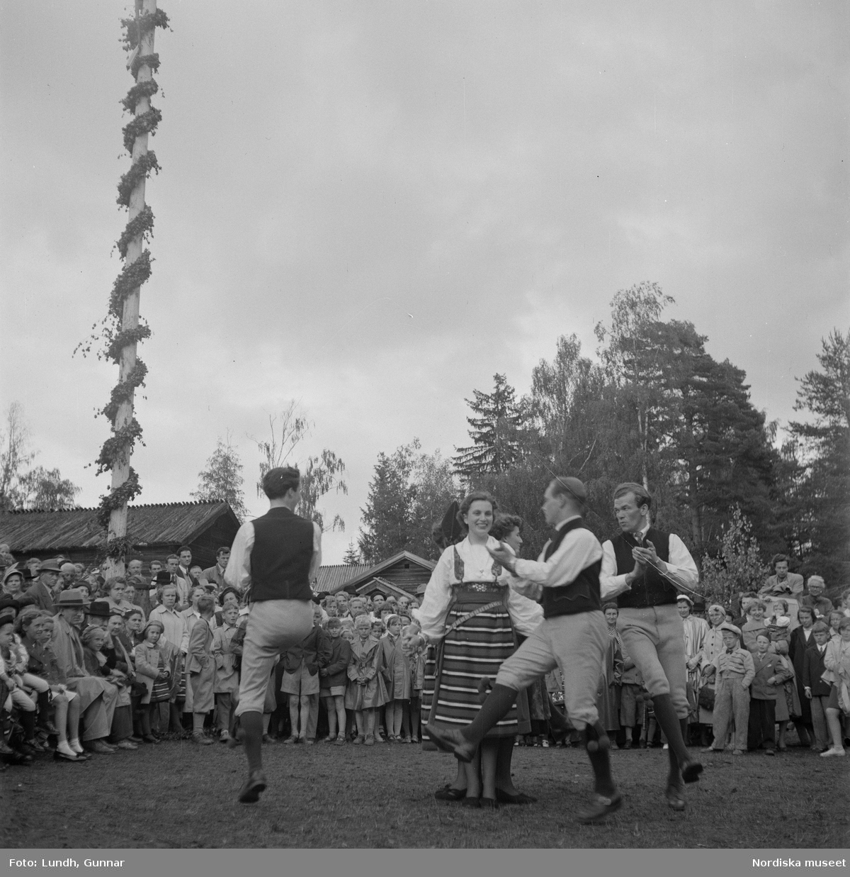 Motiv: (ingen anteckning) ;
Kvinnor och män i folkdräkt dansar vid en midsommarstång inför en publik vid midsommarfirande, tre hästskulpturer 2Jemt-Olofs hästar" (träsnidaren Jemt Olov Persson), porträtt av en man som står vid en husvägg, porträtt av en kvinna som står på en balkong.