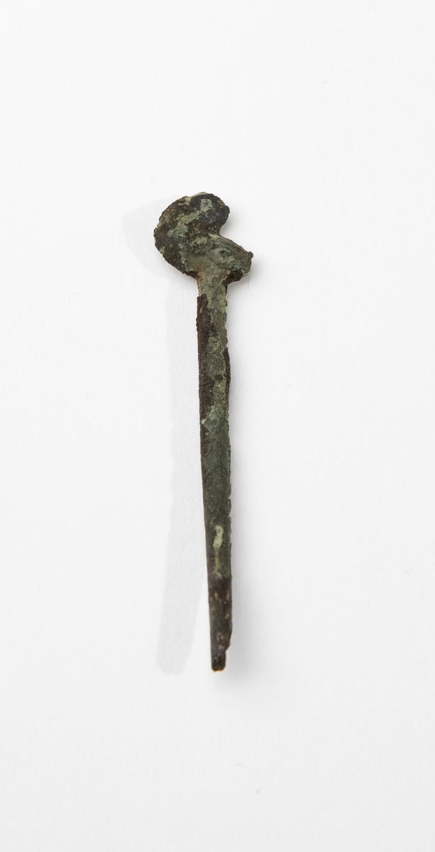 Dräktnål av brons med asymmetriskt huvud och avbruten spets. Utplattat deformerat huvud, ursprungligen runt? Påträffad i grav A25. Nålen liknar vagt så kallade fågelnålar, men denna saknar i så fall huvudet.