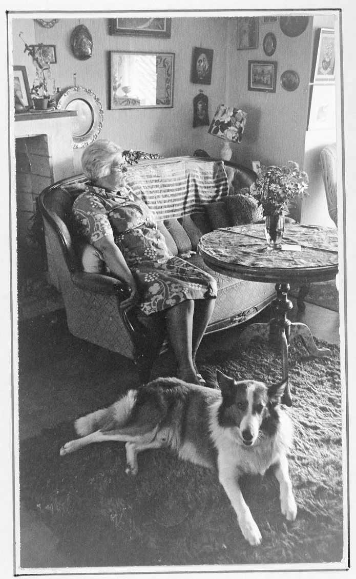 Interiör hos Karin och Bernt Rendahl. Karin sitter i hitre änden av en soffa och tittar bort mot ett fönster. Deras hund ligger på mattan vid hennes fötter och tittar mot fotografen.