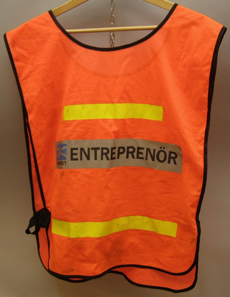 Varselväst i starkt orange färg, med reflexremsor påsydda på flera ställen. Med svart tryckt text på ryggen står vilken funktion personen som bär västen har: "Entreprenör".