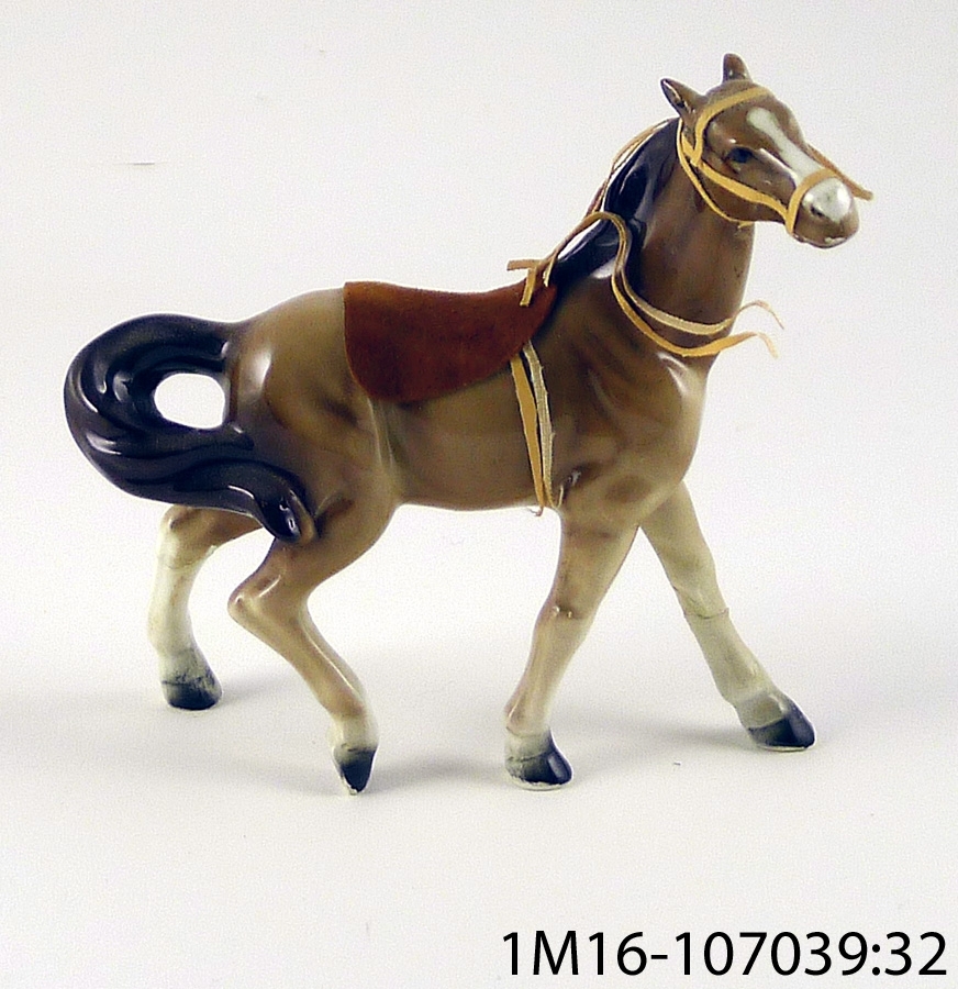 Häst i keramik, brun med sadel stigbyglar och remtyg. Hästen trasig, lagad med klister. Ena stigbygeln har lossnat.