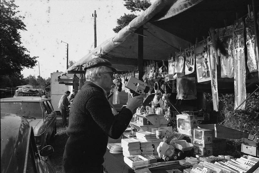 Marknad i Adelöv. En man med keps och fleecetröja står och filmar mot ett marknadsbord med leksaker.