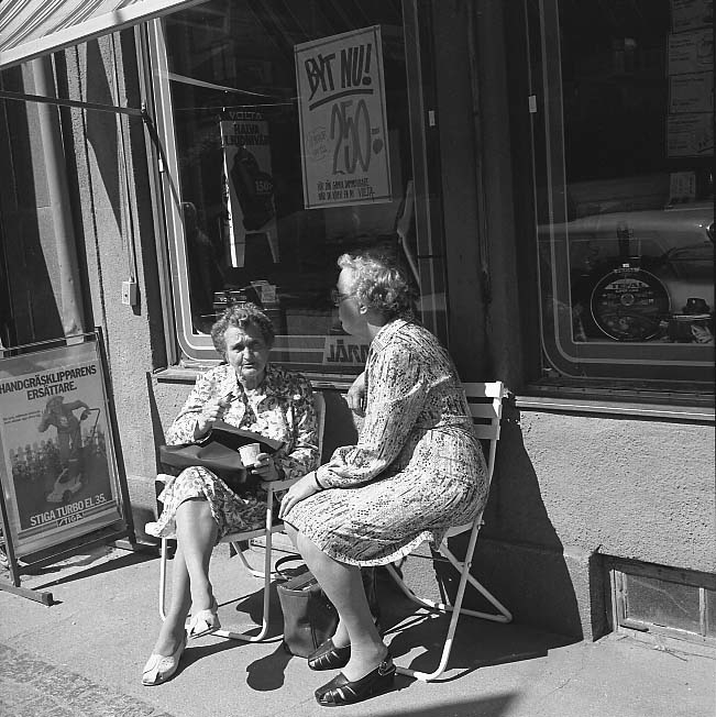 Två kvinnor i mönstrade klänningar sitter i varsin stol utanför skyltfönstret till Palm Järn på Brahegatan i Gränna. De sitter delvis i skugga.