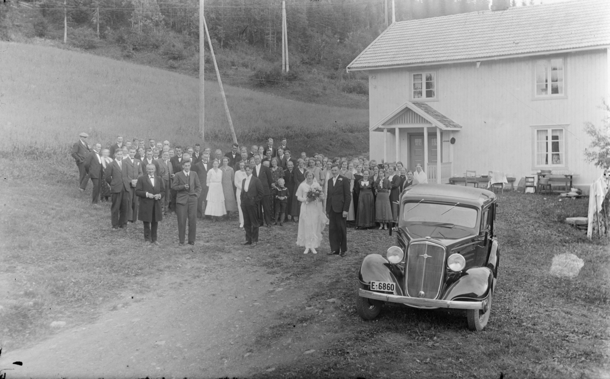 Bryllupsfeiring med brudeparet og mange gjester utenfor en bygning. En bil i forgrunnen med bilnummer E-6860.