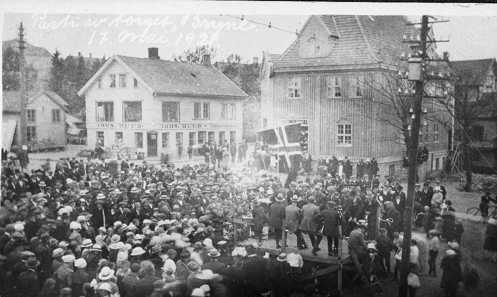 Prospektkort frå Torget på Bryne 17. mai 1921. Til v. forretningen til Johs. Ruud og til høgre Svein Tjemsland sin.