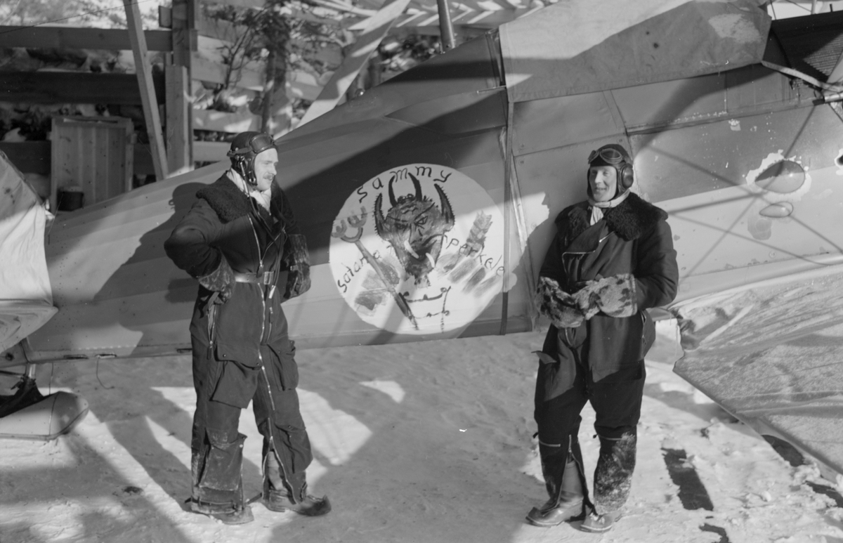 Två flygdräktsklädda flygskyttar tillhörande Svenska frivilligkåren i Finland, F 19, står framför flygplan Gloster Gladiator märkt med emblem djävulsfiguren Sammy och texten 'Satana Perkele'. Till vänster Matti Sundsten, till höger Thure Hansson.