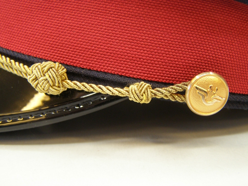 Skärmmössa med lågt kapell av vintertyg och rött mössband.
Guldfärgad stormträns, guldfärgade (1991) mösstränsknappar, samt mössmärke av 1991 års modell.