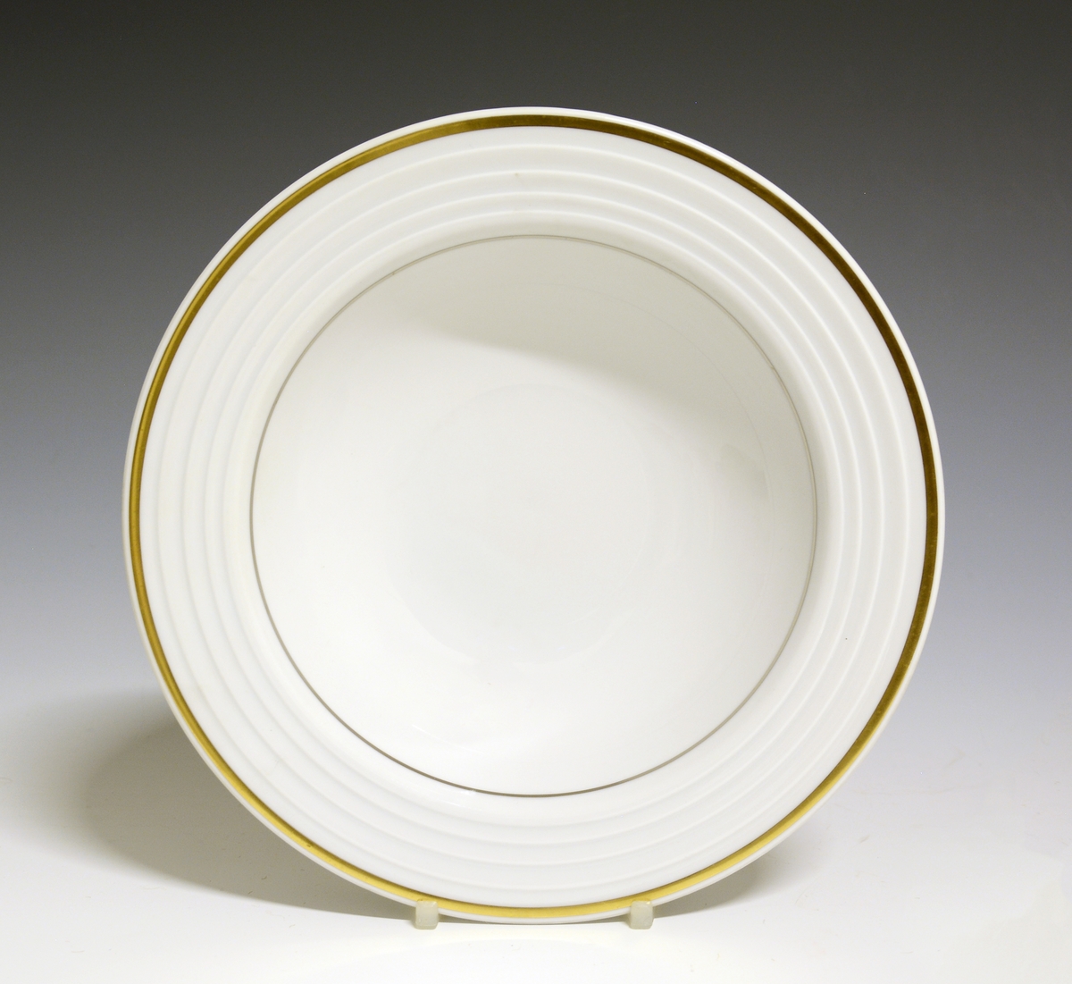 Dyp tallerken av porselen med hvit glasur. I fanens gods riller. En bredere gullstripe ytterst på fanen og en smalere innerst.
Modell: Saturn
Dekor: Comet