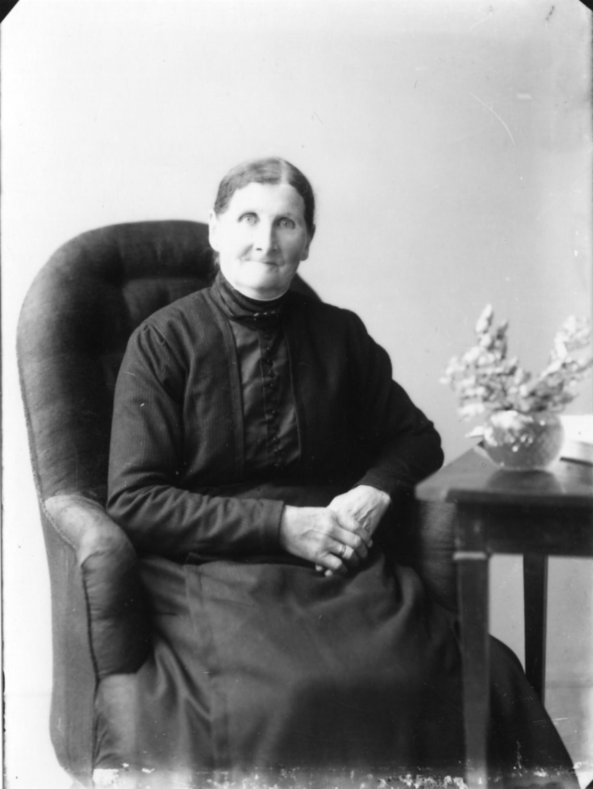 Porträtt av en äldre kvinna i mörk blus och kjol som sitter i en fåtölj vid ett bord med en blomvas. Hennes namn är Maria Johansson, från Bohult.