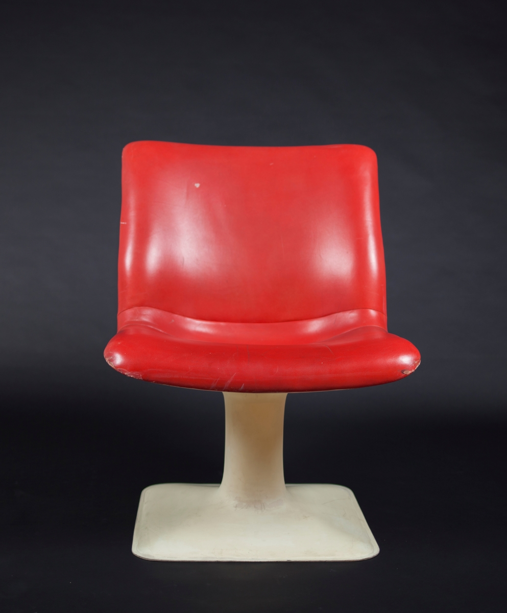 Glassfiberstol støpt i to deler; nedre del med kvadratisk fotstykke og midtsøyle og øvre del med sammenhengende sete og rygg. Øvre del er trukket med rødt skinn.