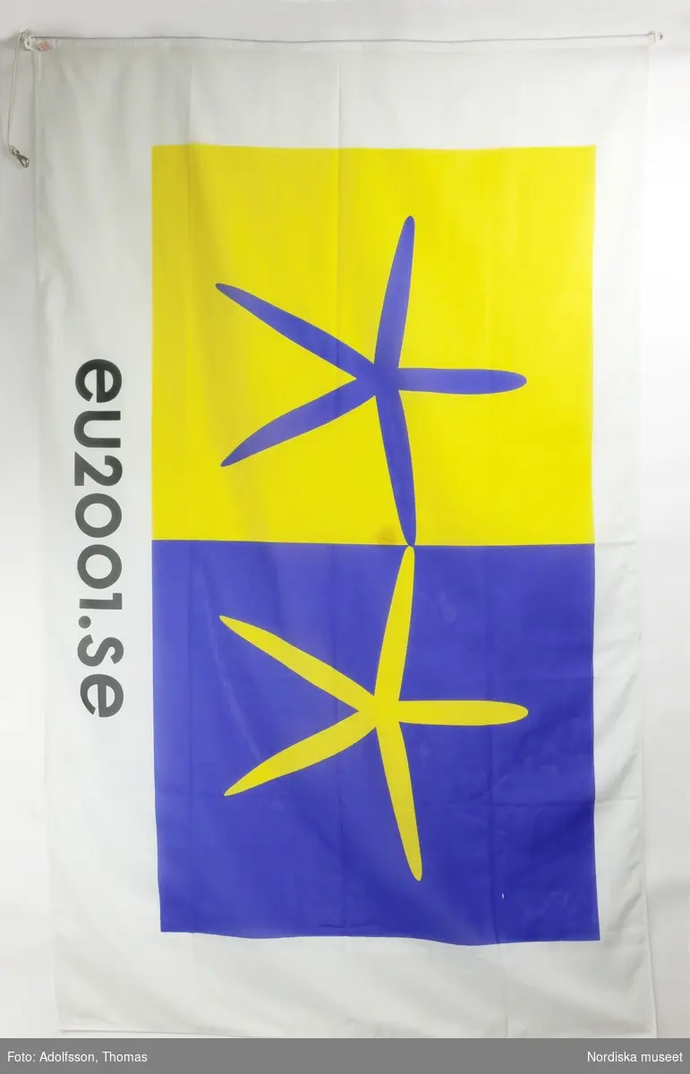 Flagga. Rektangulär flagga av polyester. På båda sidor tryckt dekor i gult och blått mot vit botten med den officiella logotypen för Sveriges ordförandeskap 2001 och text ”eu2001.se” i svart. Maskinfållad. Ena kortsidan förstärkt med ett band, i hörnen flaggkrokar av vit plast, den ena med rep och karbinhake av vitmetall. Vidsydd lapp med texten ”FLAGGFABRIKEN KRONAN 100% POLYESTER 042/200540 HELSINGBORG SWEDEN” samt tvättanvisning.

Vid mitten en fläck.

L  241,5 cm
Br 147 cm