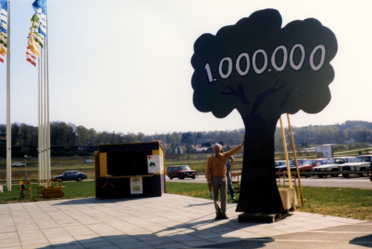 Röda Korsets insamling "En miljon träd" på Ikeas parkeringsplats år 1985. Alve Brandin (1923 - 2001) står och lutar sig mot det konstgjorda trädet. I bakgrunden till vänster ser man Ikeas flaggor.