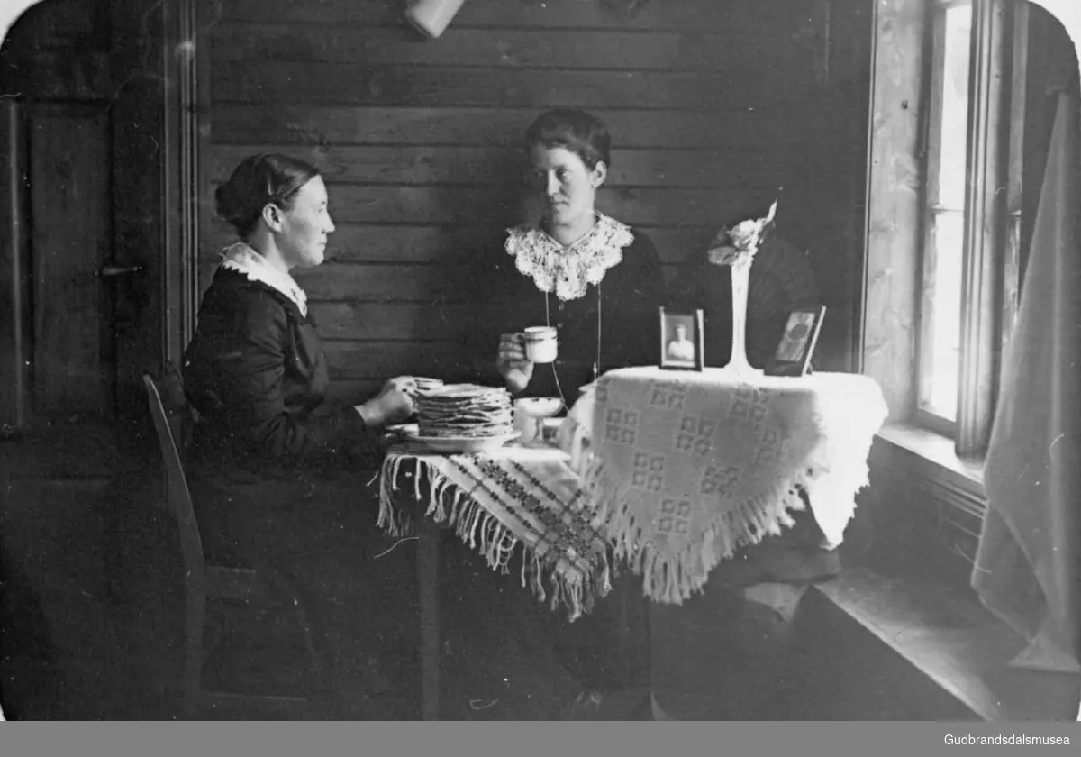 To kvinner sitter ved bordet og drikker kaffe og konverserer, bordet er dekket med diverse; servise, tallerken med skrivarbrød?

