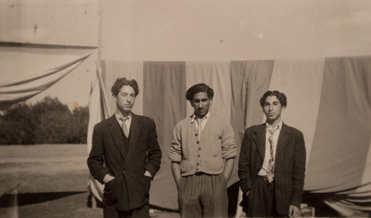 Tre unga romska män framför en upphängd tältduk. Två bär kavaj medan en tredje i mitten bär kofta.