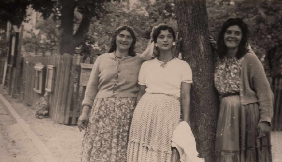 Tre romska kvinnor på Storgatan i Storvik. I bakgrunden syns ett staket och anslagstavlor. Bilden är tagen en sommardag i juli 1950.