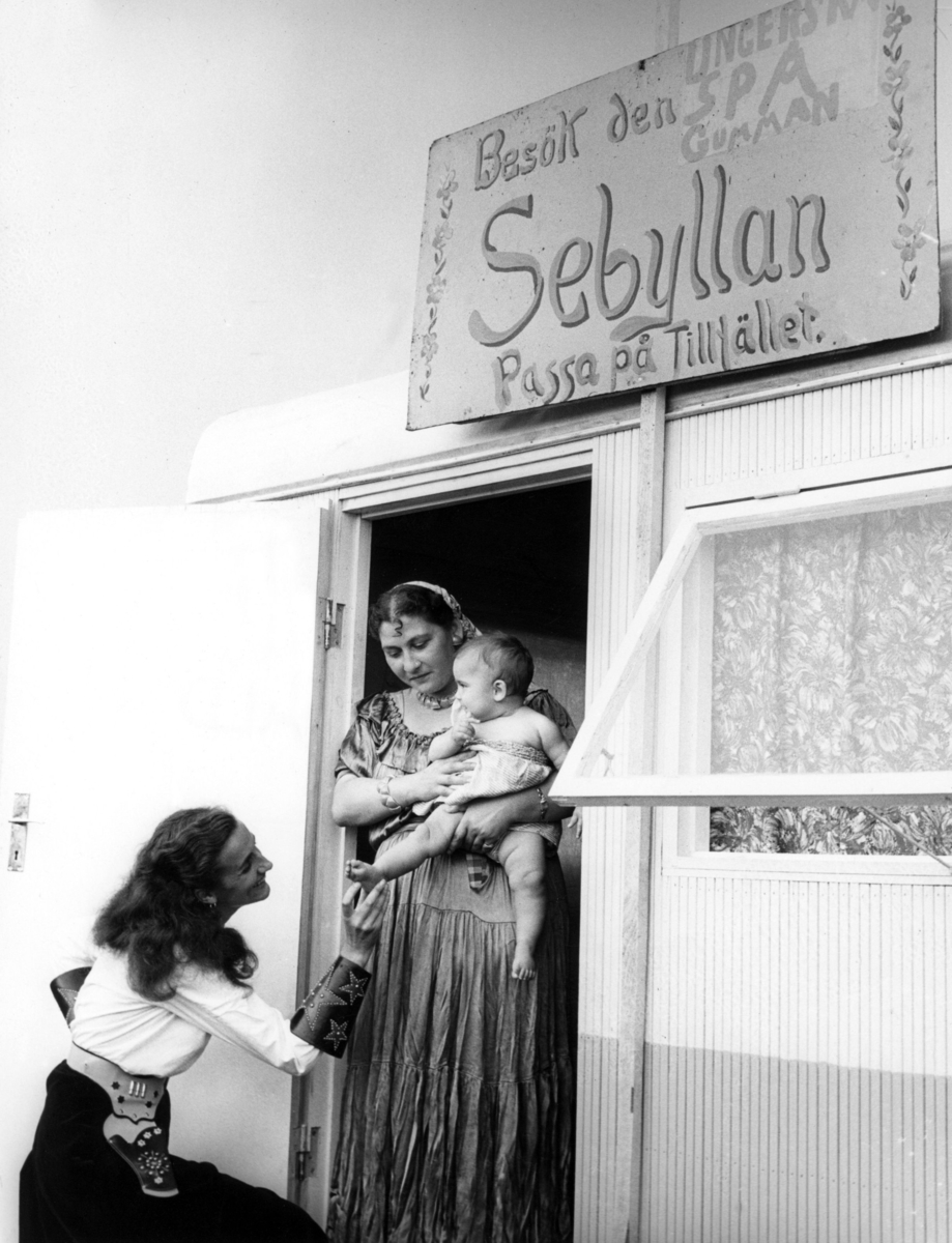 Spåkvinna utanför sin vagn på Kiviks marknad 1957. På skylten står "Besök den underbara spågumman 
Sebyllan. Passa på tillfället". Marknader har ofta varit samlingsplatser för romer.
