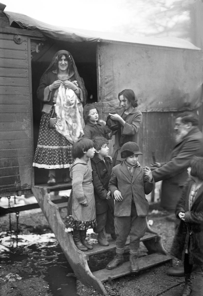En romsk familj står på och runt trappan till en bostadsvagn. Högt upp på trappan står en kvinna som bär en dikhlo. Runt henne finns sex barn i olika åldrar.