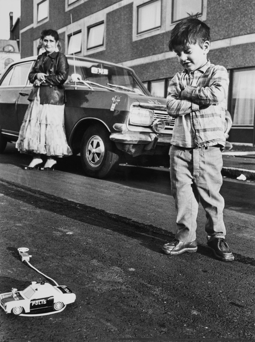 En pojke står på en gata i Göteborg och tittar på den leksaksbil han fått av socialförvaltningen. I bakgrunden syns en romsk kvinna stå lutad mot en bil.