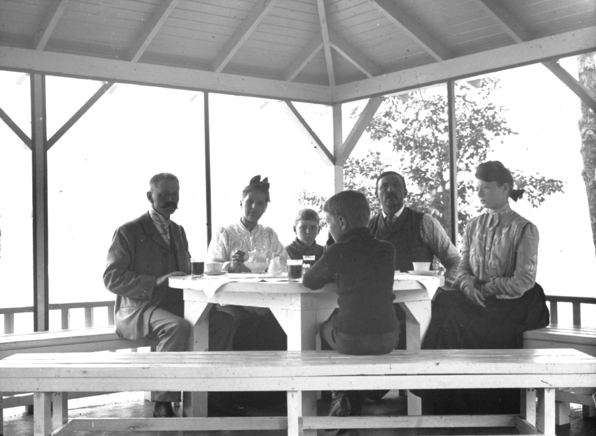 Seks personer rundt et bord i et lysthus.