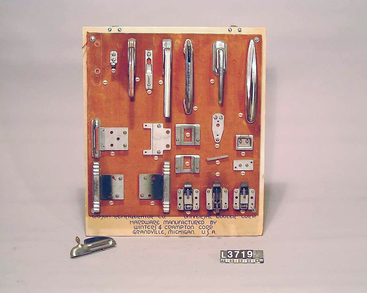 Gångjärn, handtag och låsannordningar till Gibsons kylskåp. Samtliga delar är uppsatta på en träplatta som är klädd med tyg.