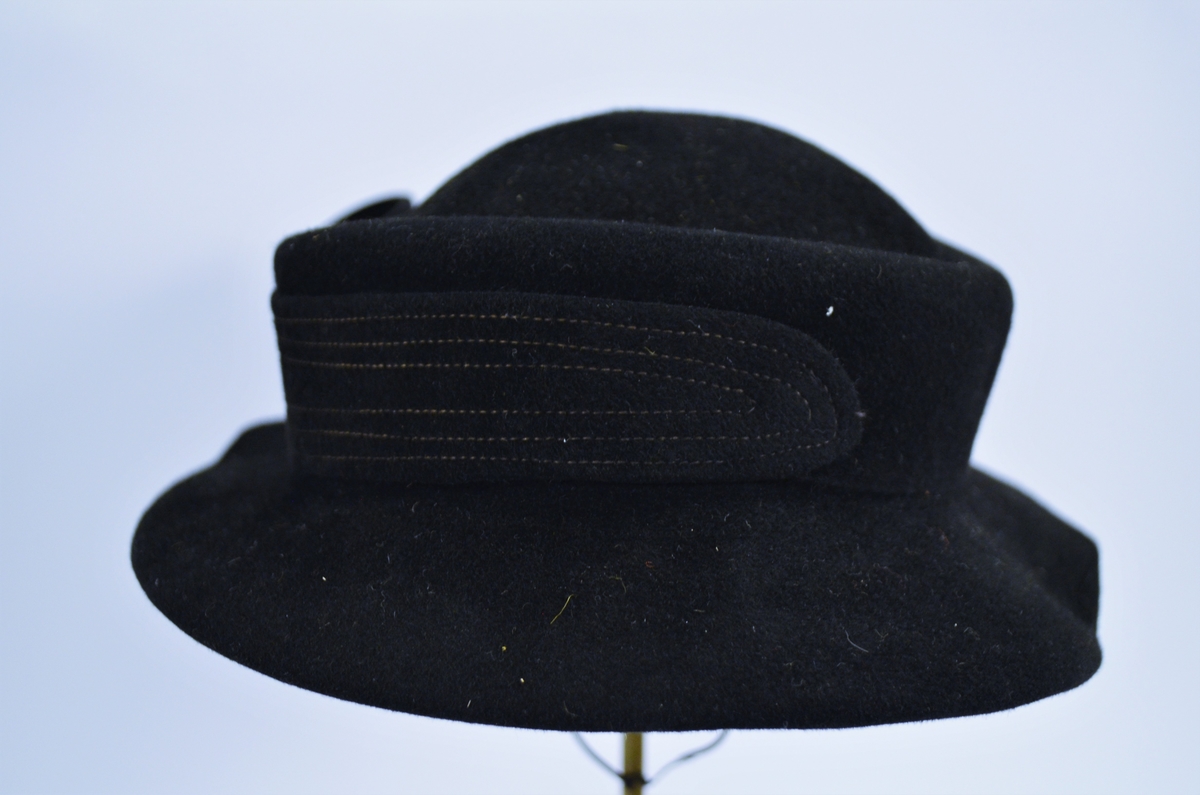 Hatt i mørk blå fløyel, pynta med blått ripsband og perler. 
Ripsband rundt kanten inni, og hattestrikk for å feste hatten.