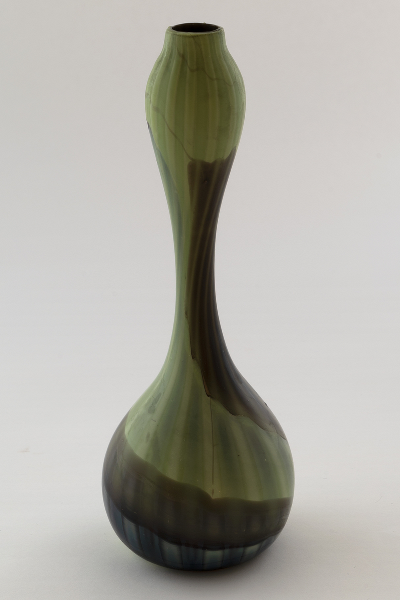 Høyreist kalebassformet vase i farvet glass. Yttersiden veksler mellom lysegrønne og brune partier med noen blålige innslag, som snor seg i en spiralbevegelse oppover korpus. Nedover vasen løper brede vertikale striper, som veksler mellom lyse og mørkere sjatteringer av bunnfargen.