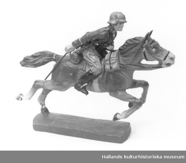 Leksakssoldat på häst, av trämassa på ståltrådsskelett i tysk uniform. Målade i grönt, brunt, svart, gult, rött, vitt och skärt.a) Höjd: 6 cm, bredd: 5 cm.