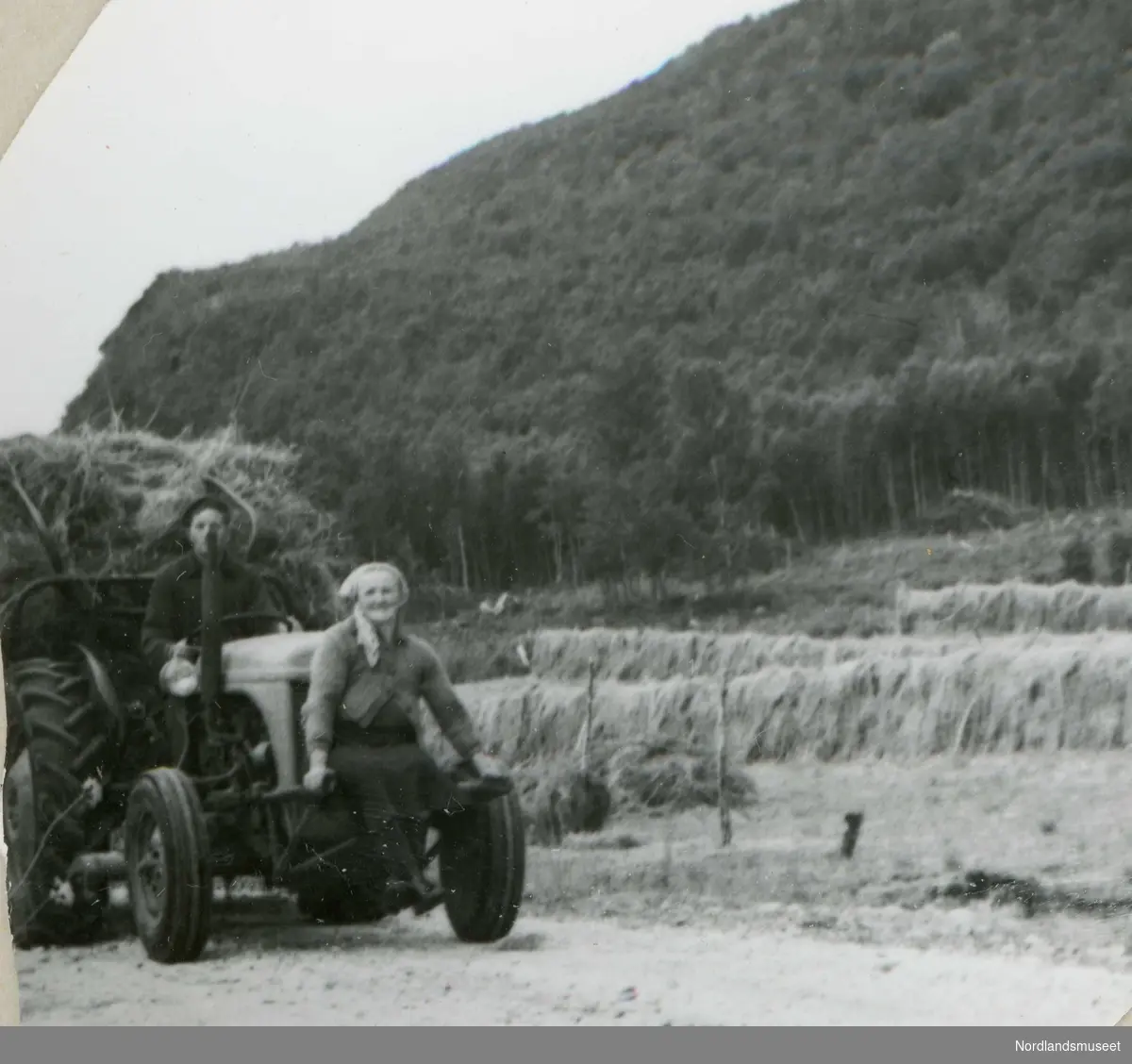 Innkjøring av tørrhøy på gården Fredvang på Holmvåg. Konrad Berntsen kjører traktor, og kona Hildur sitter foran på ballast-ramma. I bakgrunnen,  hesjer.