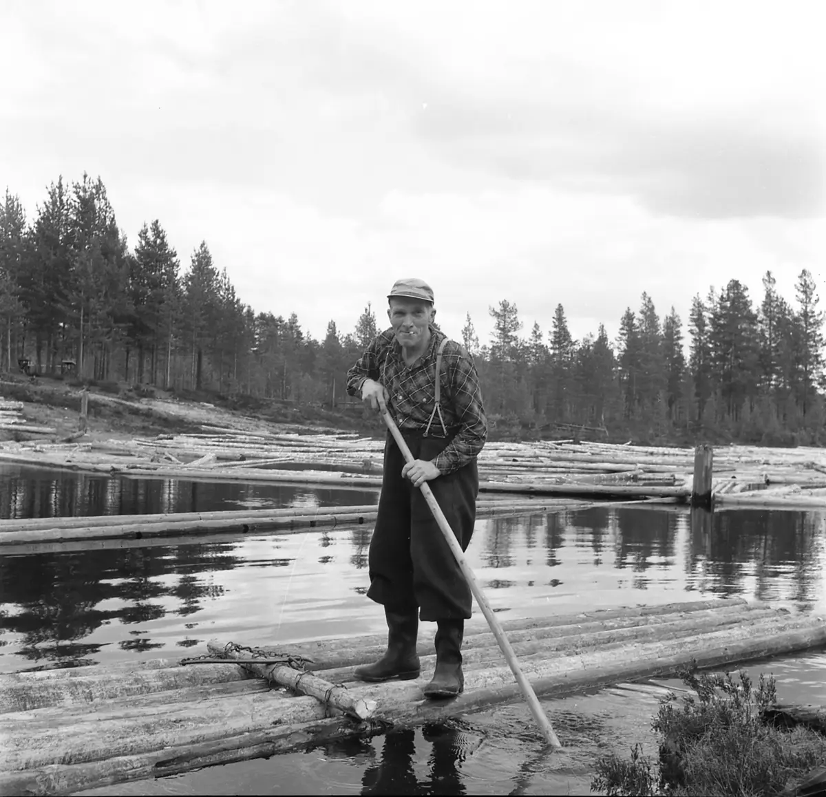 ​Fløtersjef Gotmar Sletengen på tømmerflåten ved Ringlidammen i Julussa. Han smiler lurt mens han ser rett inn i kamera, med sigaretten hengende i munnviken.