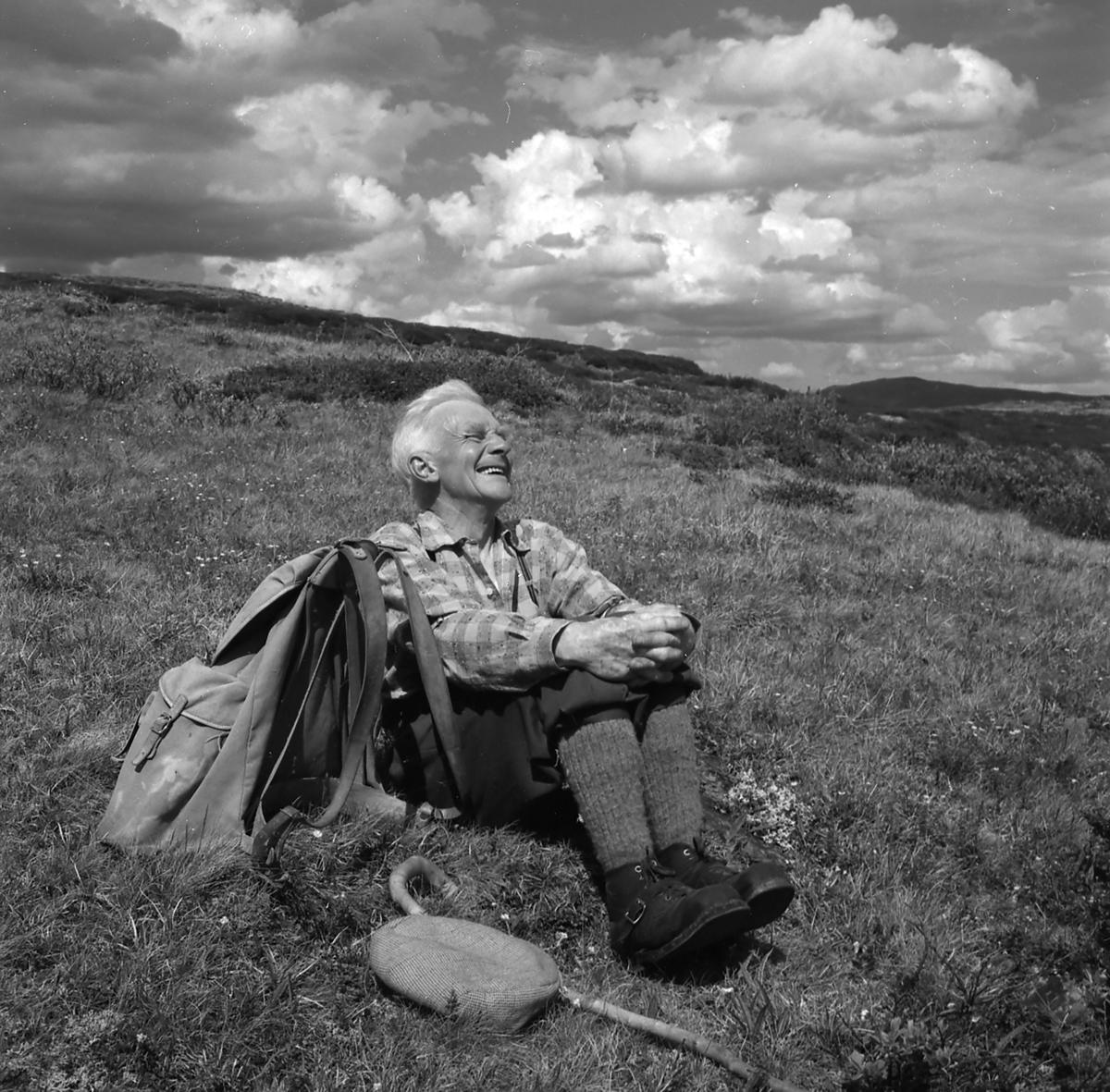 ​En eldre man tar en pause i fjellet. Han sitter på bakken, smiler mot sola og har ryggsekken stående ved siden av seg. Fra hesteslepp på Bekkelegeret i Folldalsfjellene, Oppdal 1960.