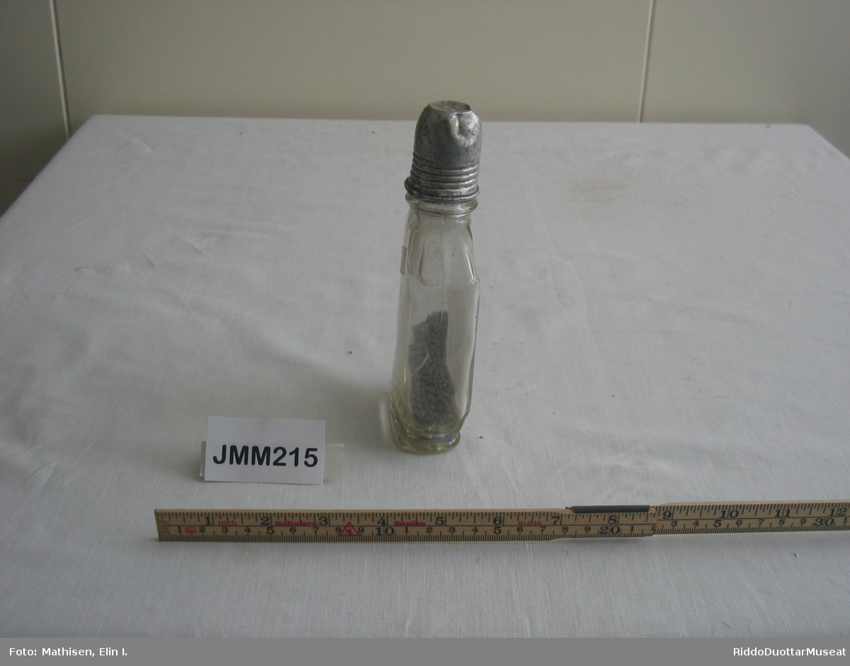 Glassflaske (lommelerke) som inneholder hagl, ammunisjon, for å lade patroner selv. Glasset i flasken er ujevn (bølget). Lokket av metall er formet som et lite beger med synlige gjenger utenpå.