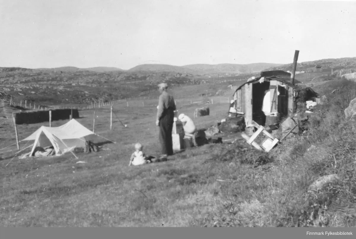 Bildet viser en mann, Georg Adriansen, og en liten hytte med pipe i Gamvik. Ved siden av Georg sitter et lite barn på bakken. Litt nedenfor dem er det slått opp et telt og lenger ned på bakken står ei hesja. Hytta var boligen de hadde den første tiden etter brannen, sommeren 1945.