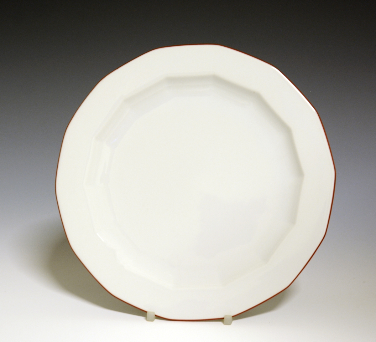 Mangekantet tallerken av porselen med hvit glasur. Dekorert med rød strek ytterst på fanen. 
Modell: Octavia, tegnet av Grete Rønning i 1977.
Dekor: Rød Strek