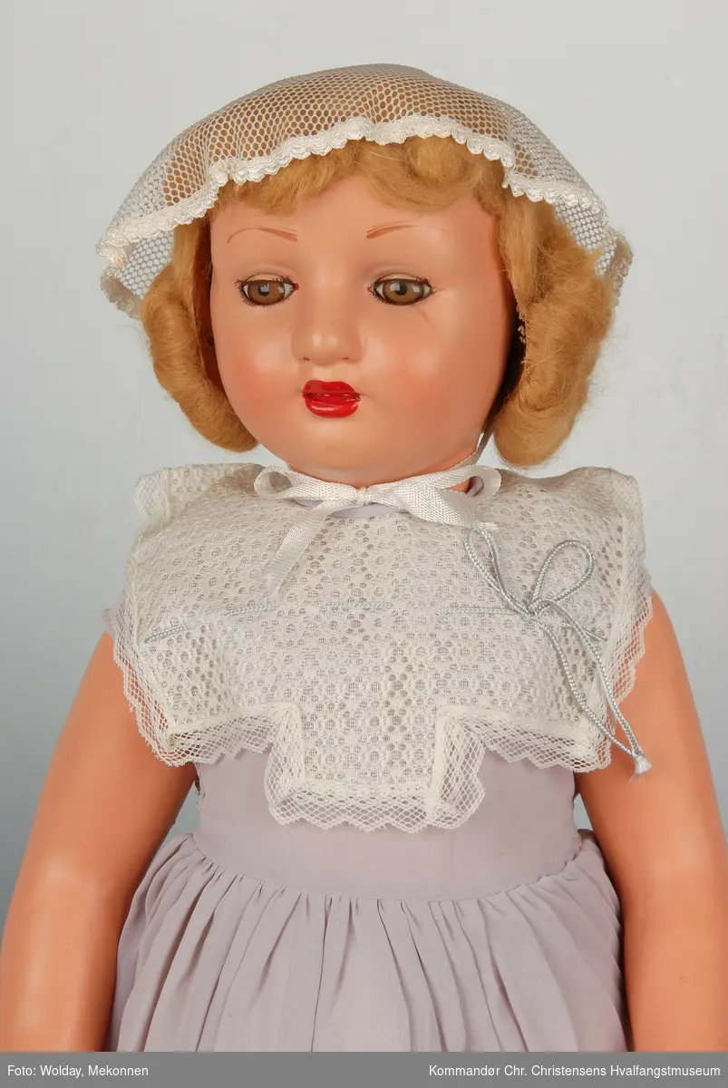 Dukke med blondt hår, kysehatt, syrinfarget kjole, hvite halvsokker og plastsandaler. 