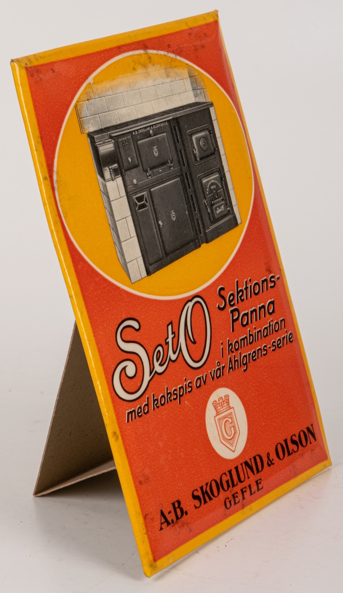 Reklamskylt för "SetO Sektionspanna i kombination med kokspis av vår Ahögrens-serie"
Plåtskylt med baksida av papp. Orange och gul med motiv av järnspis.