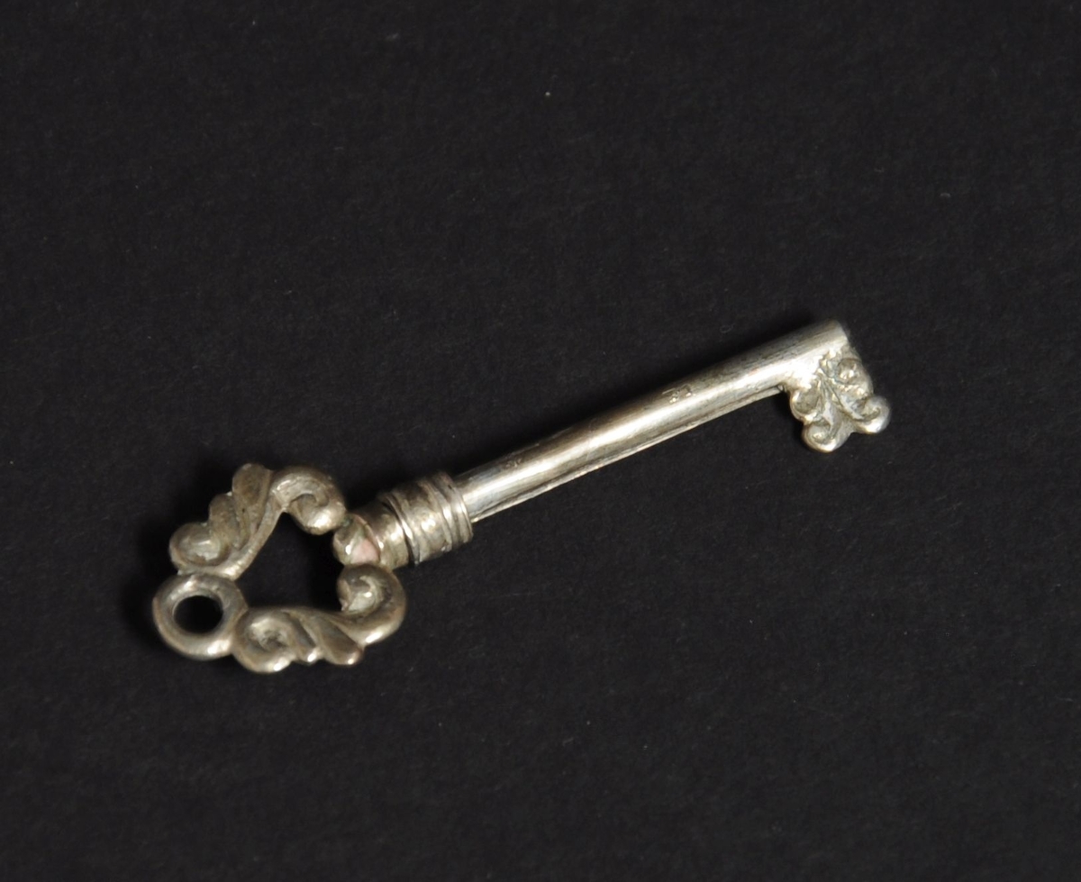 Neglerensker av sølv. Forestiller nøkkel, med nagle som kan skyves ut fra tuppen med et skyveelement bak på nøkkelen. Naglen har spiralprofil innerst.