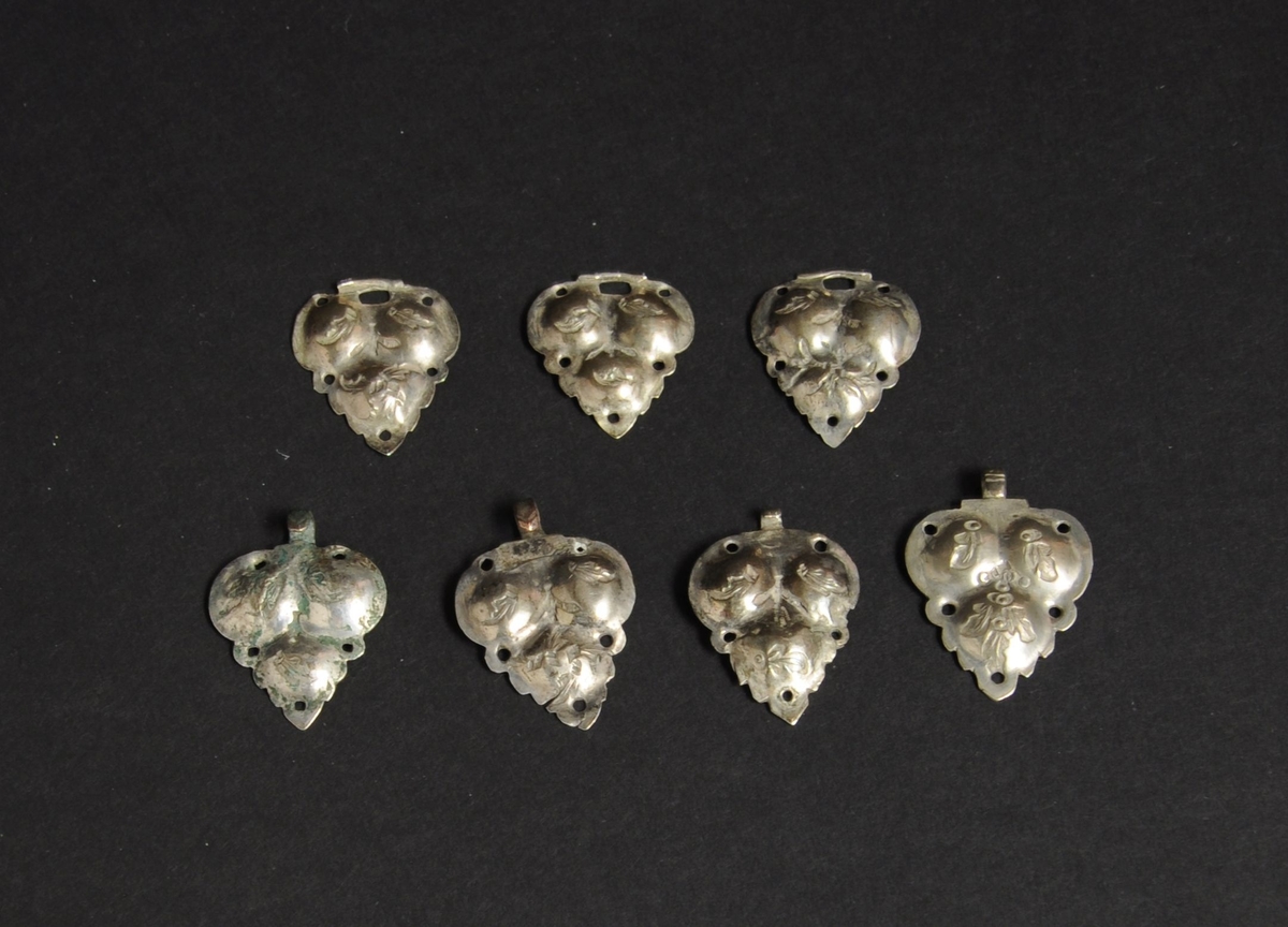 7 halvdeler til halspenner, tre par og en ekstra del. Halsspennene er av sølv og har gravert dekor. Spennene har bladform, med fem gjennombrutte hull for tekstilfeste.