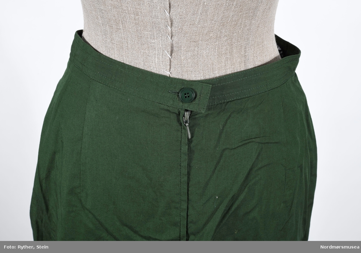 Grønt skjørt som festes med knapp og glidelås. Spesielt utformet snitt foran, som et foldeskjørt - overlappende åpent snitt fra linning og ned.