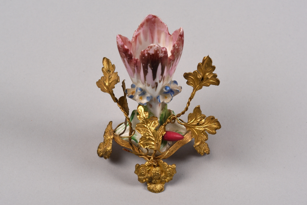 Porslinsvas med ställning i guldmålad metall. Vasen är formad som en blomsterkalk med dekor av platsiskt gjorda blommor och blad i rokokostil. Vasen står i en ställning i form av blad med tvinnade stjälkar. De fyra fötterna är i utformade som sköldar.