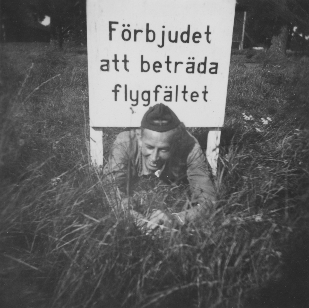 Värnpliktig man vid F 3 Malmen ligger på mage under en skylt med texten "Förbjudet att beträda flygfältet" vid flygdagen Flygets dag på F 3 Malmen, 1938.