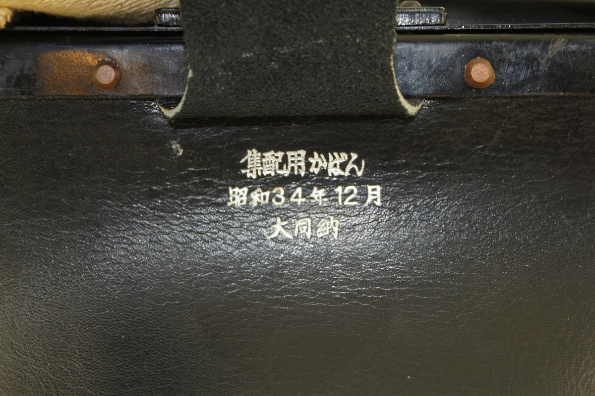 Brevbärarväska av svart läder med bygel, tillslutningsanordning och axelrem, 4 cm bred av naturfärgad väv. På framsidan ett påtryckt "silver" emblem och under bygelplösen en japansk inskription.

Ej använd.