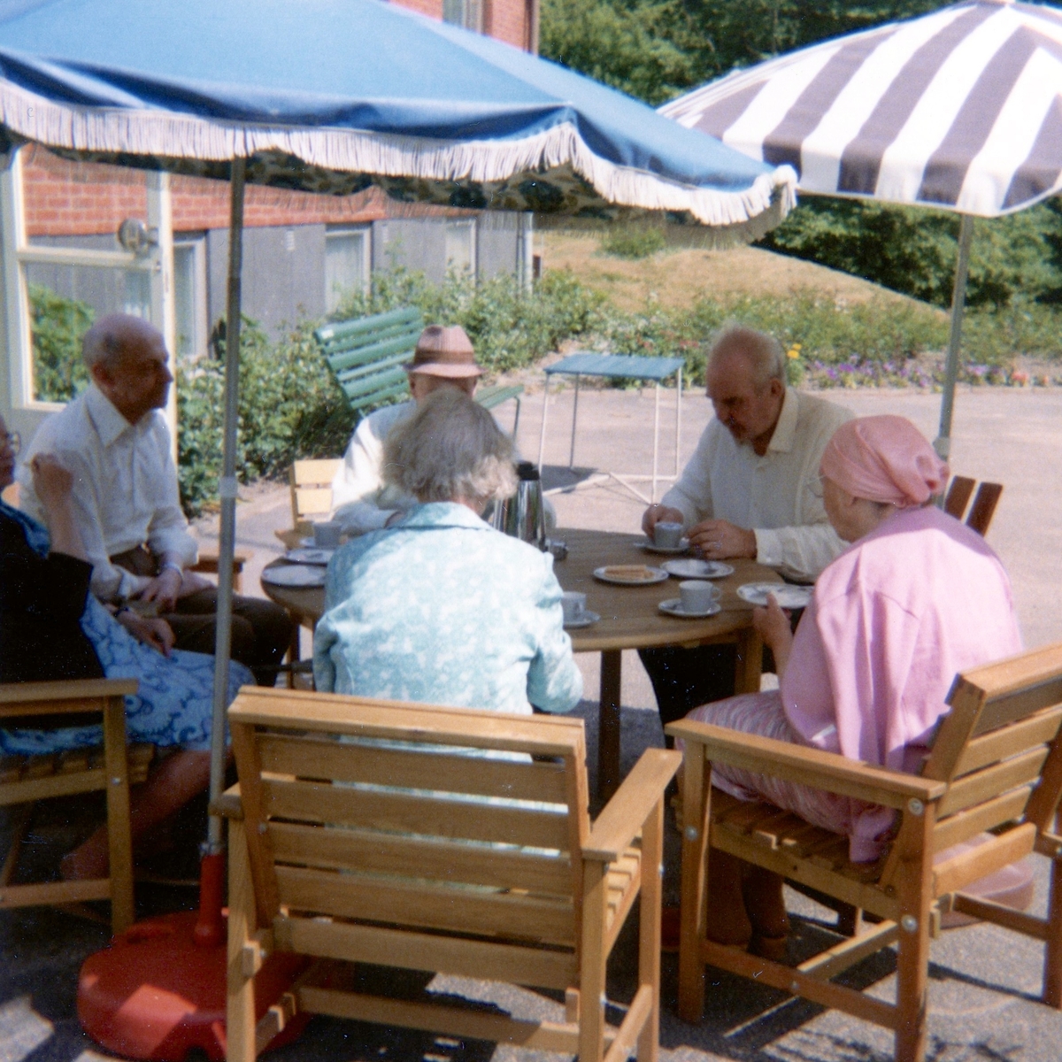 Sex personer intar midsommarkaffe, sittandes under parasoller utanför Brattåshemmet 1980. Namnuppgifter saknas.