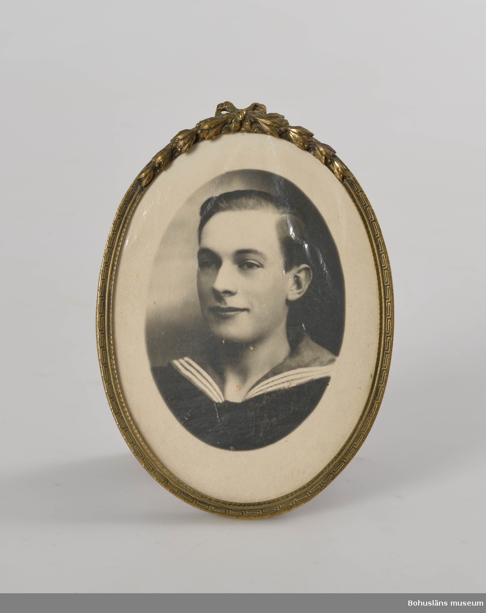 Svartvitt fotografi inramat i oval fotoram av gulmetall. 
Motiv: Carl Gustaf Bernhardson i sjömanskrage.