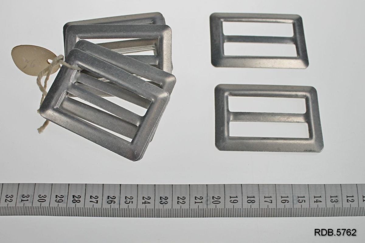 En bunt med 17 sølvfargede beltespenner i aluminium. Rektangelformet med midtstolpe. Sammenbundet med hyssing med oval prislapp i papp.