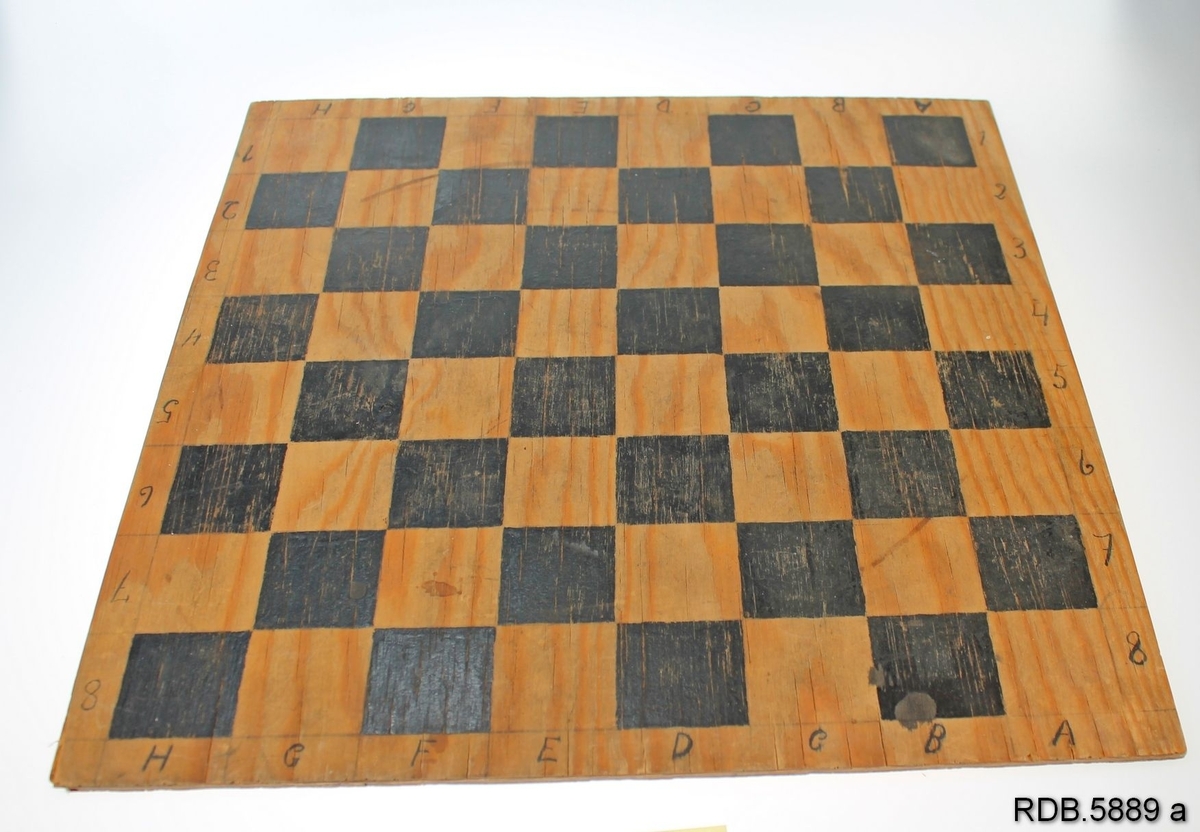 Hjemmelaget sjakkbrett skåret av ei finèrplate. Plata er oppruta med blyant og påmalt svarte kvadratiske ruter. Langs kanten på to sider er påskrevet for hånd bokstaver fra A-H og på de to andre sidene tall fra 1-8.